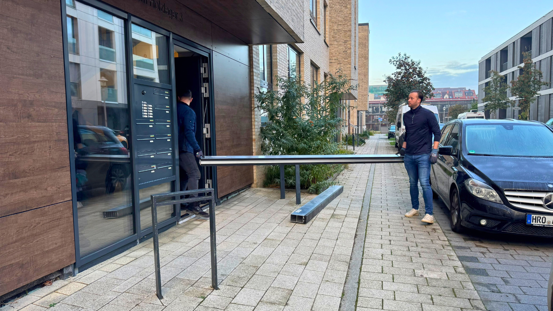 Zwei Männer vor einem Gebäude mit einer Alustange in den Händen.